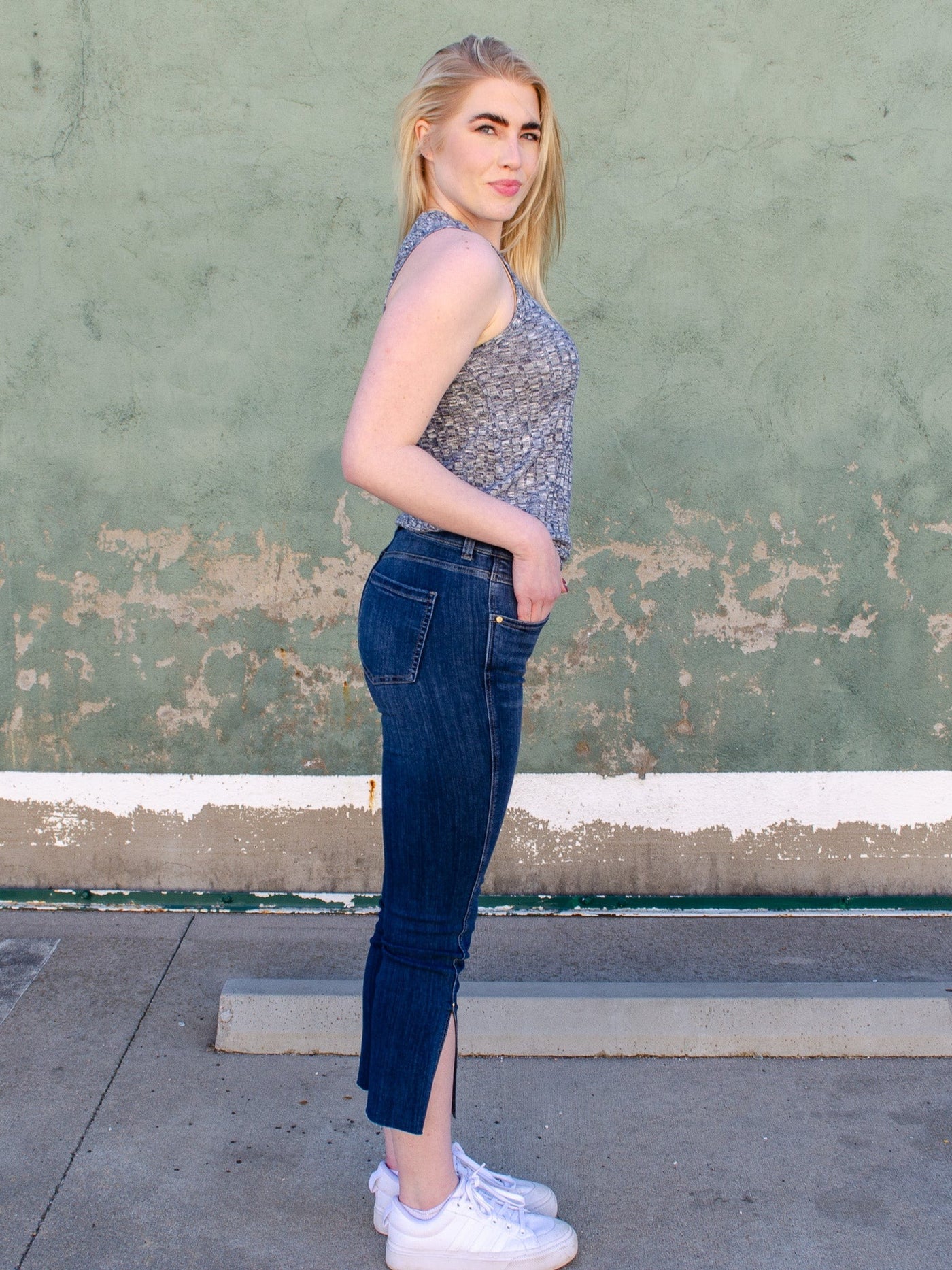 Model is wearing a dark wash 3/4th length fitted jean with a slit on each ankle. Jeans are paired with a grey tank top. 