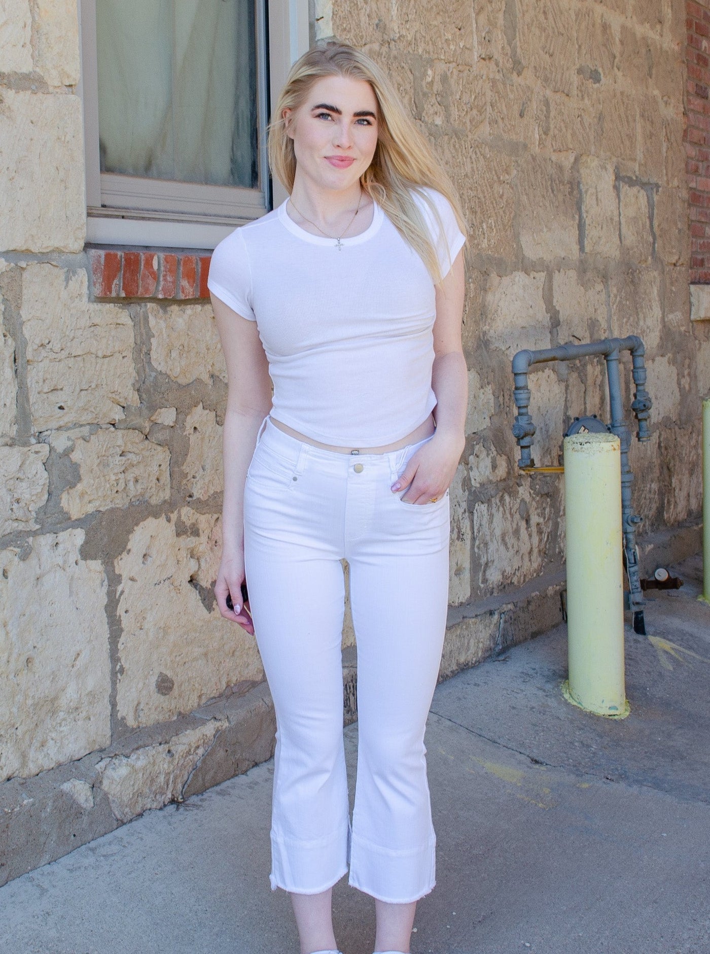 Model is wearing white mid rise denim jeans with frayed details at the hemming of the ankle. Jeans are worn with a white shirt. 