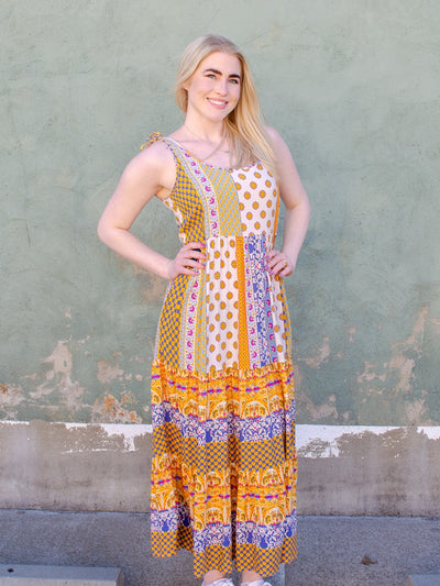 Model is wearing a multi colored multi print flowy maxi dress with a scoop neck line and tie details on the straps. 