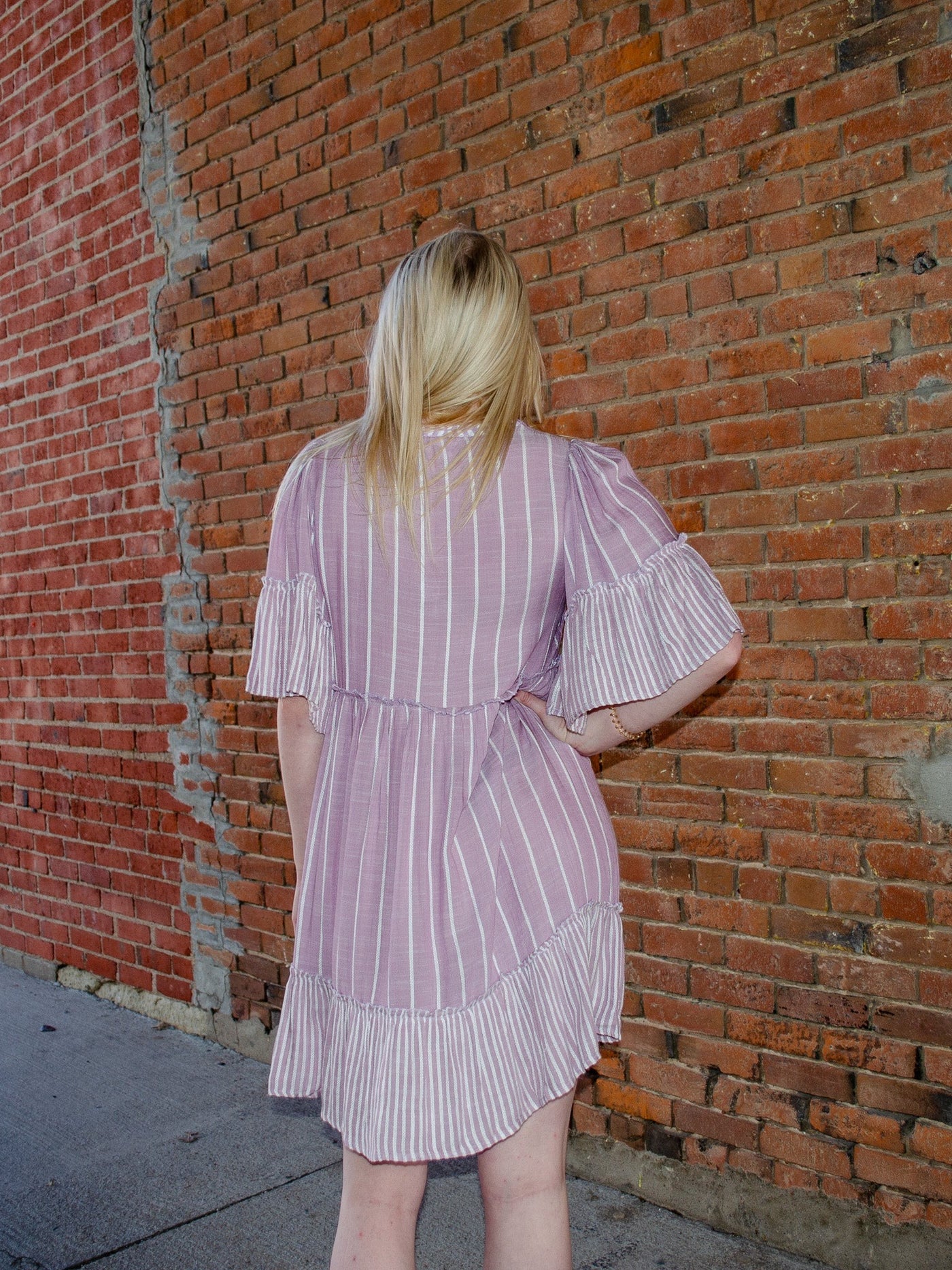 Model is wearing a striped purple and white half sleeve dress that is thigh length and cinches at waist. The neckline has string details to allows for adjusting of the open neck.