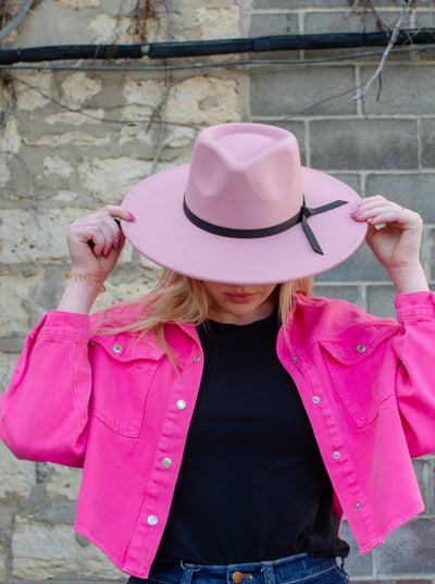 Model is wearing a wide brimmed fedora style hat thats light pink and has a faux leather black strap.