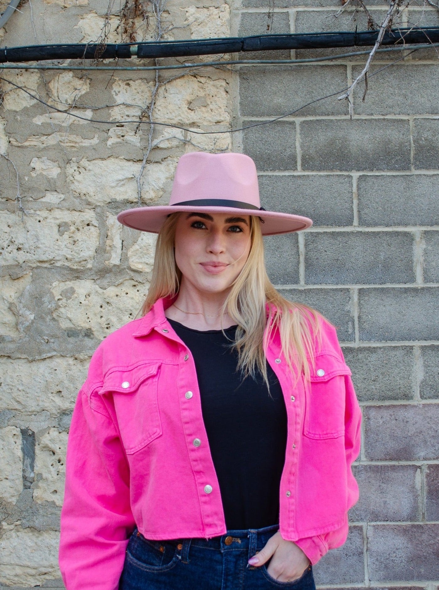 Model is wearing a wide brimmed fedora style hat thats light pink and has a faux leather black strap.