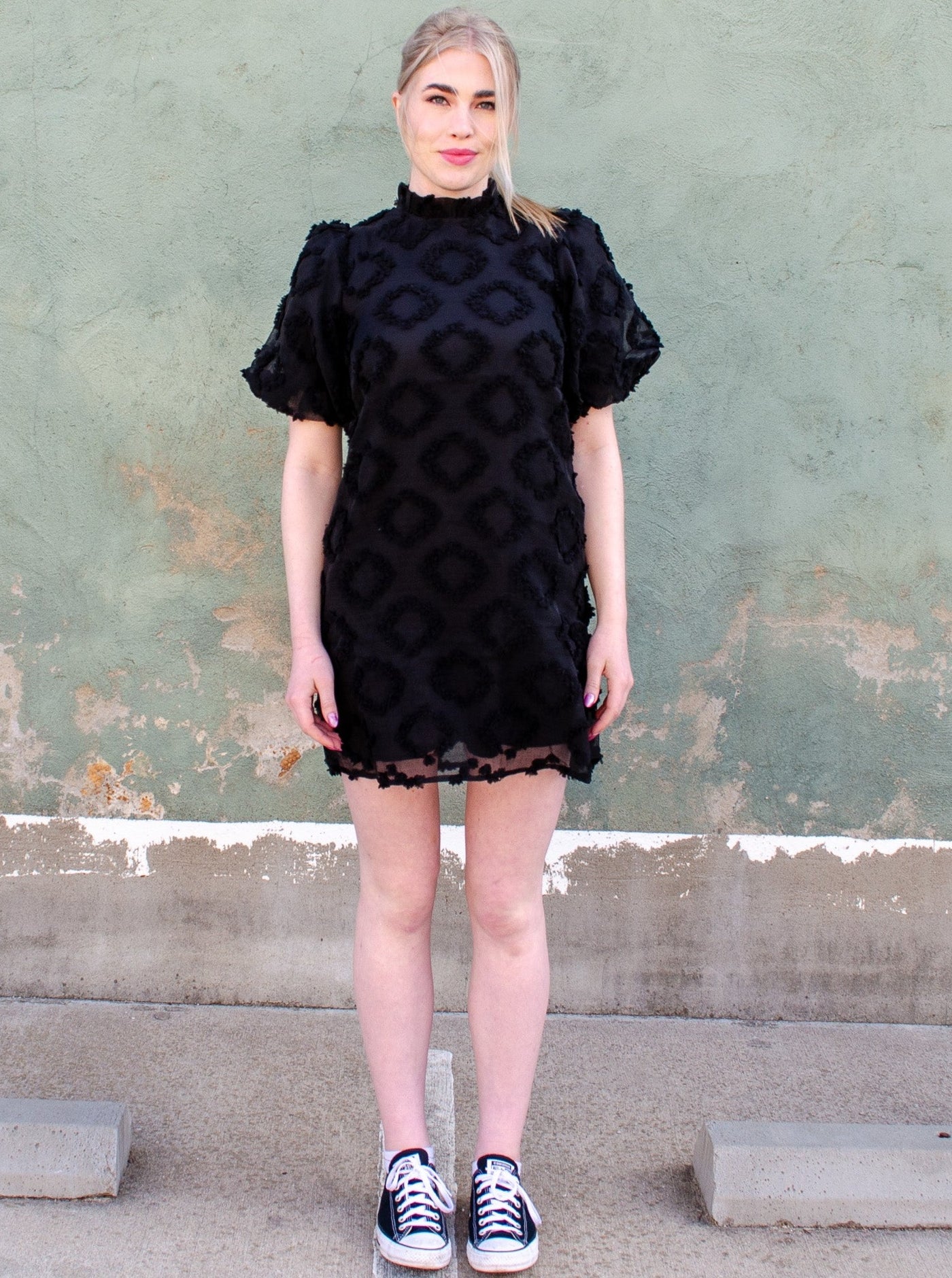 Model is wearing a textured puffed sleeve mock neck black midi dress. Dress is worn with converse tennis shoes. 