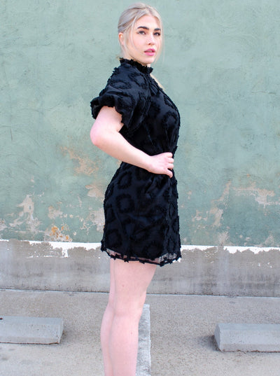 Model is wearing a textured puffed sleeve mock neck black midi dress. Dress is worn with converse tennis shoes. 