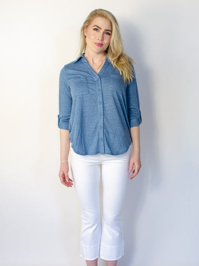 Model is wearing a button up collared half sleeve blouse. Blouse is a denim blue color and worn with white pants.