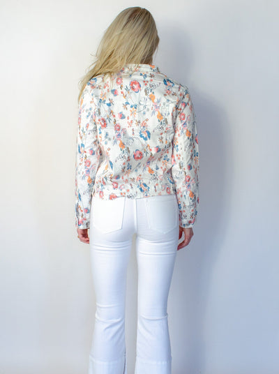 Model is wearing a white denim jean jacket with floral printed detail. Jean jacket is worn with white jeans.