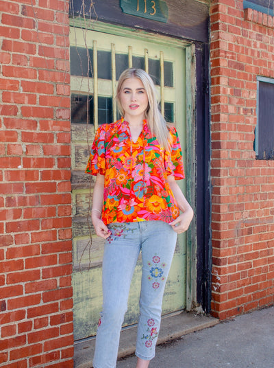 Model is wearing a red, orange, blue, and green floral printed split neck blouse. Blouse has a tie detail at the neck. 
