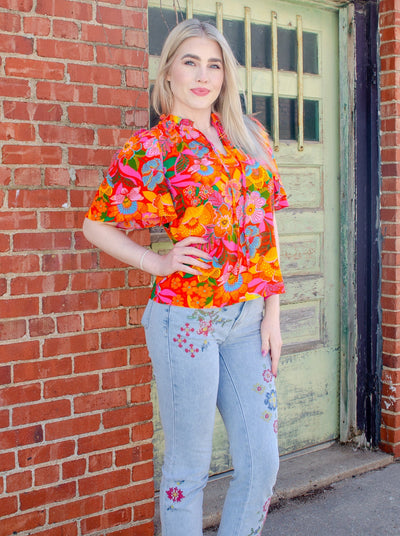 Model is wearing a red, orange, blue, and green floral printed split neck blouse. Blouse has a tie detail at the neck. 