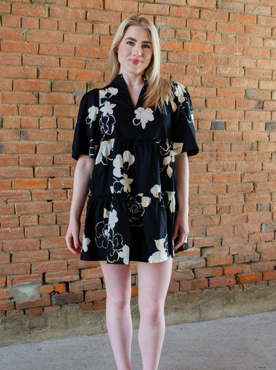 Model is wearing a baby doll style black dress with white and beige floral print. Dress is short sleeve and has a v neck. Dress hits mid thigh. 