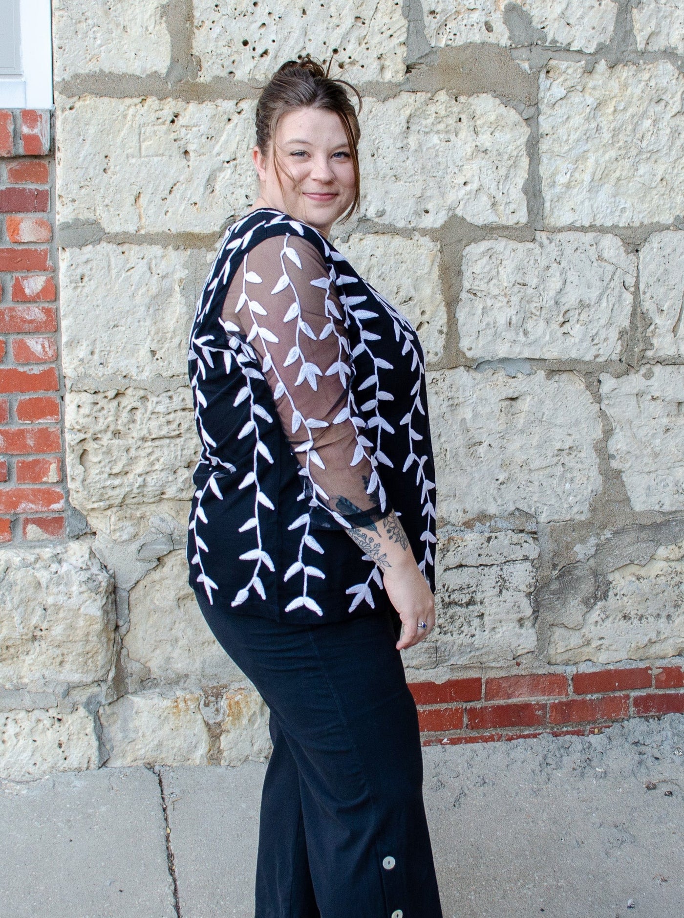 Model is wearing a black and white embroidered long sleeve with sheer fabric on the sleeves. Top is worn with black pants.