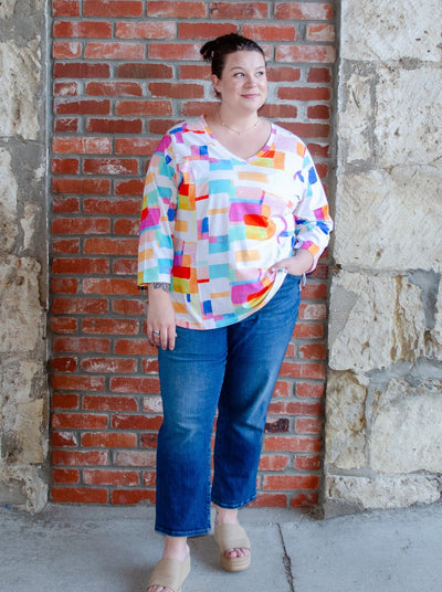 Model is wearing a V-neck style white long sleeve with multi color block print. Worn with dark wash blue jeans.