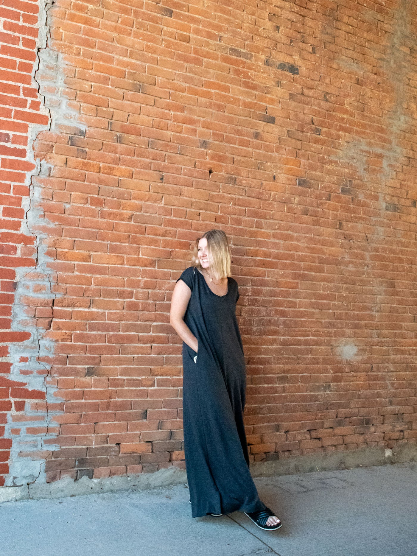 A model wearing an oversized, wide leg charcoal jumpsuit. The jumpsuit features a scoop neck and cap sleeve. The model has it paired with a black sandal.