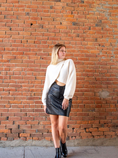 A model wearing a black pleather skirt with seam details. The model has it paired with a cream turtleneck sweater and black booties.