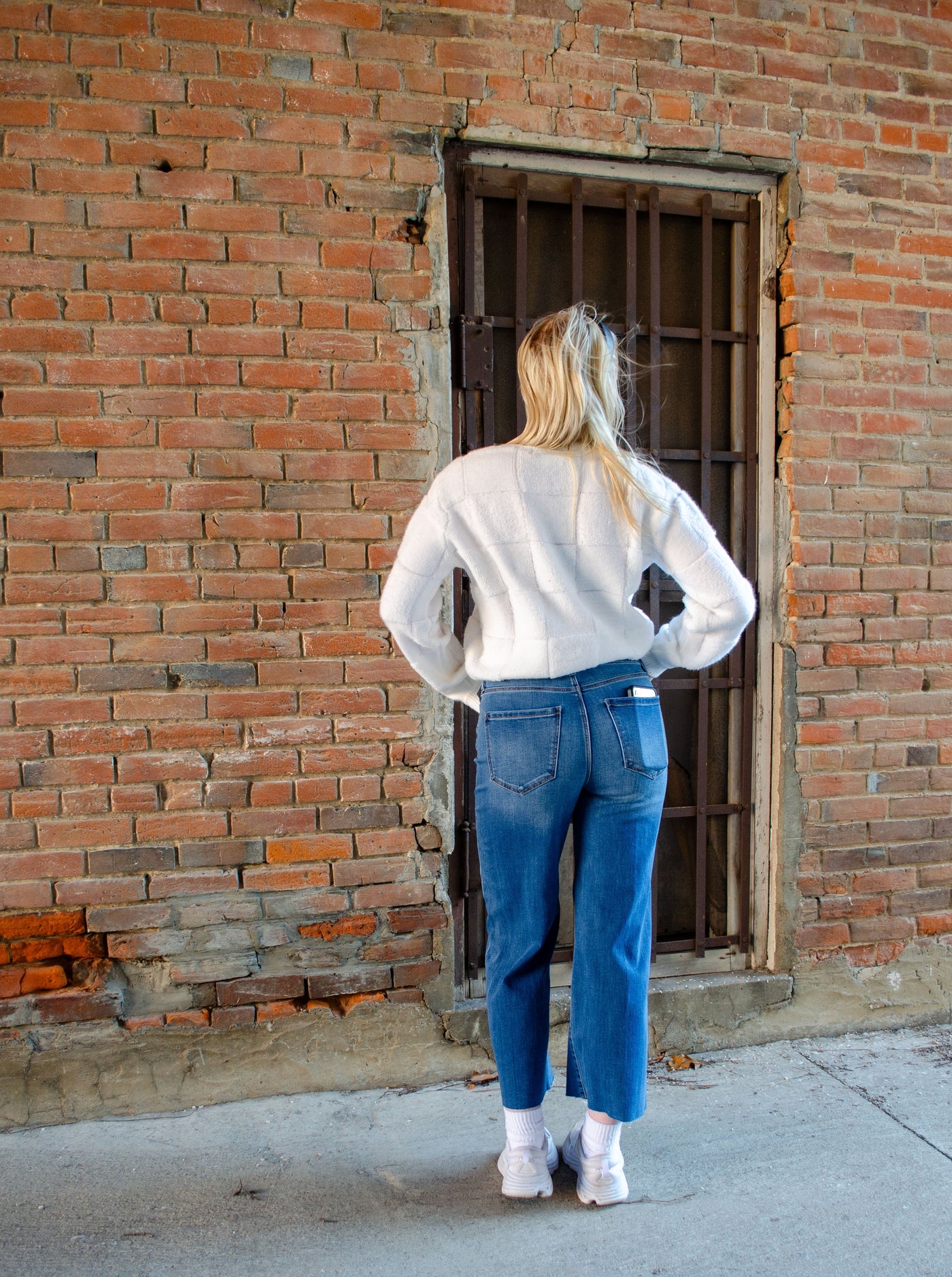 Model wearing a cream textured sweater with checkered details. Sweater crops at waist and is paired with blue jeans.