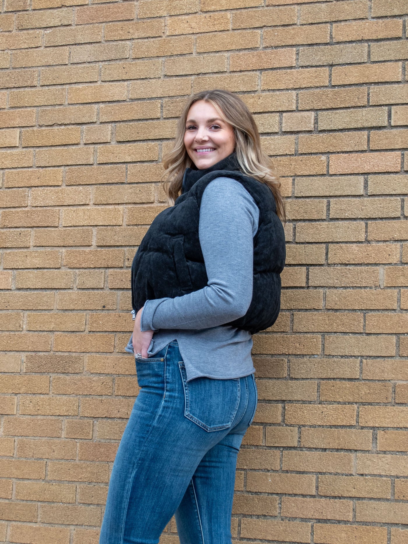 A model wearing a black corduroy puffer vest. The model has it paired with a gray v-neck and medium wash jeans.