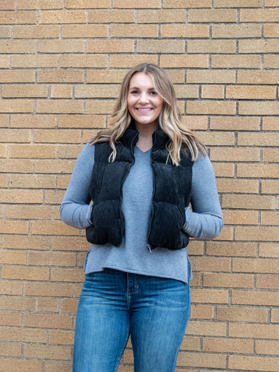 A model wearing a black corduroy puffer vest. The model has it paired with a gray v-neck and medium wash jeans.