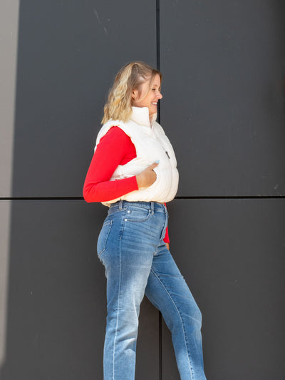 A model wearing a cream, cropped puffer vest. The model has it paired with a read mock neck underneath and light wash jeans.