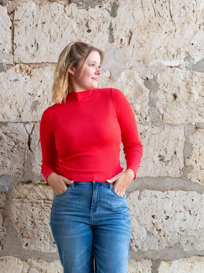 A model wearing a red, ribbed mock neck sweater top. She has it paired with a medium wash jean.