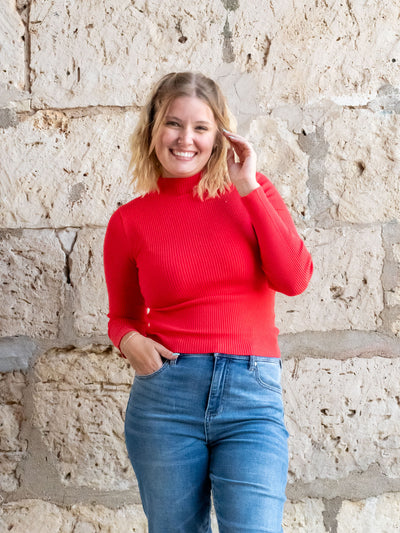 A model wearing a red, ribbed mock neck sweater top. She has it paired with a medium wash jean.