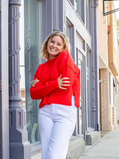 A model wearing a red sweater with hem and sleeve details and side slits. She has it paired with white jeans.