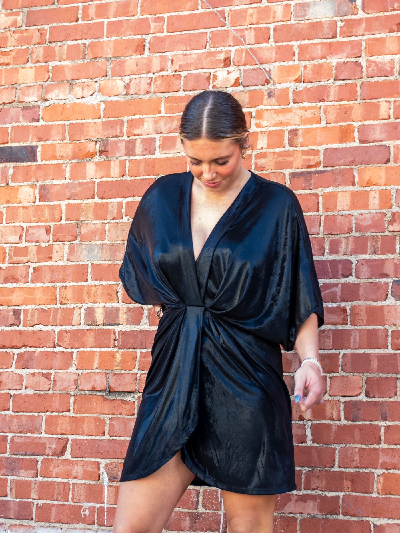 A model wearing a black mini dress with a deep v-neck, half dolman sleeve, and shiny finish.