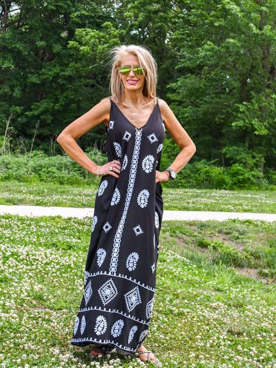 A model wearing a black v-neck maxi dress with embroidered details, spaghetti straps, and side pockets. She has it on with sandals.