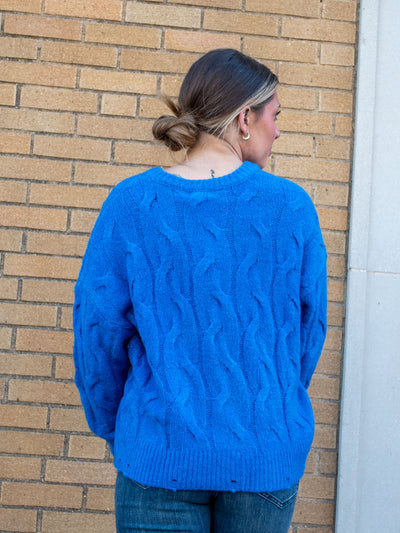 A model wearing a blue cable knit sweater with distressing on the hem. The model has it paired with a dark wash jean.