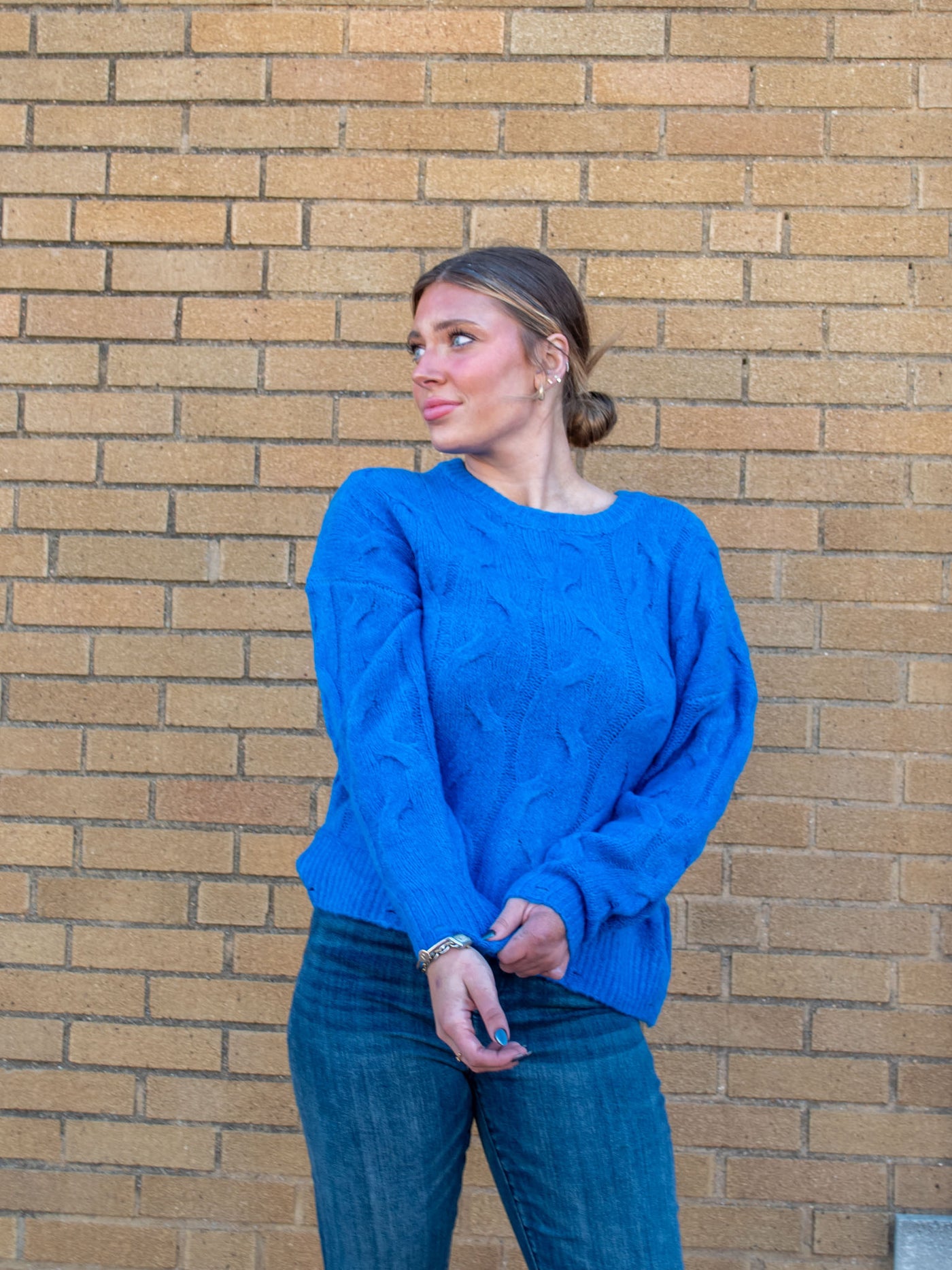 A model wearing a blue cable knit sweater with distressing on the hem. The model has it paired with a dark wash jean.