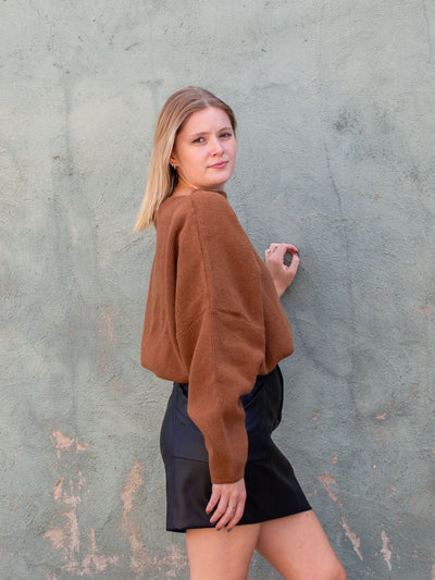 A model wearing a brown, off the shoulder slouchy sweater. The model has it paired with a black pleather skirt.