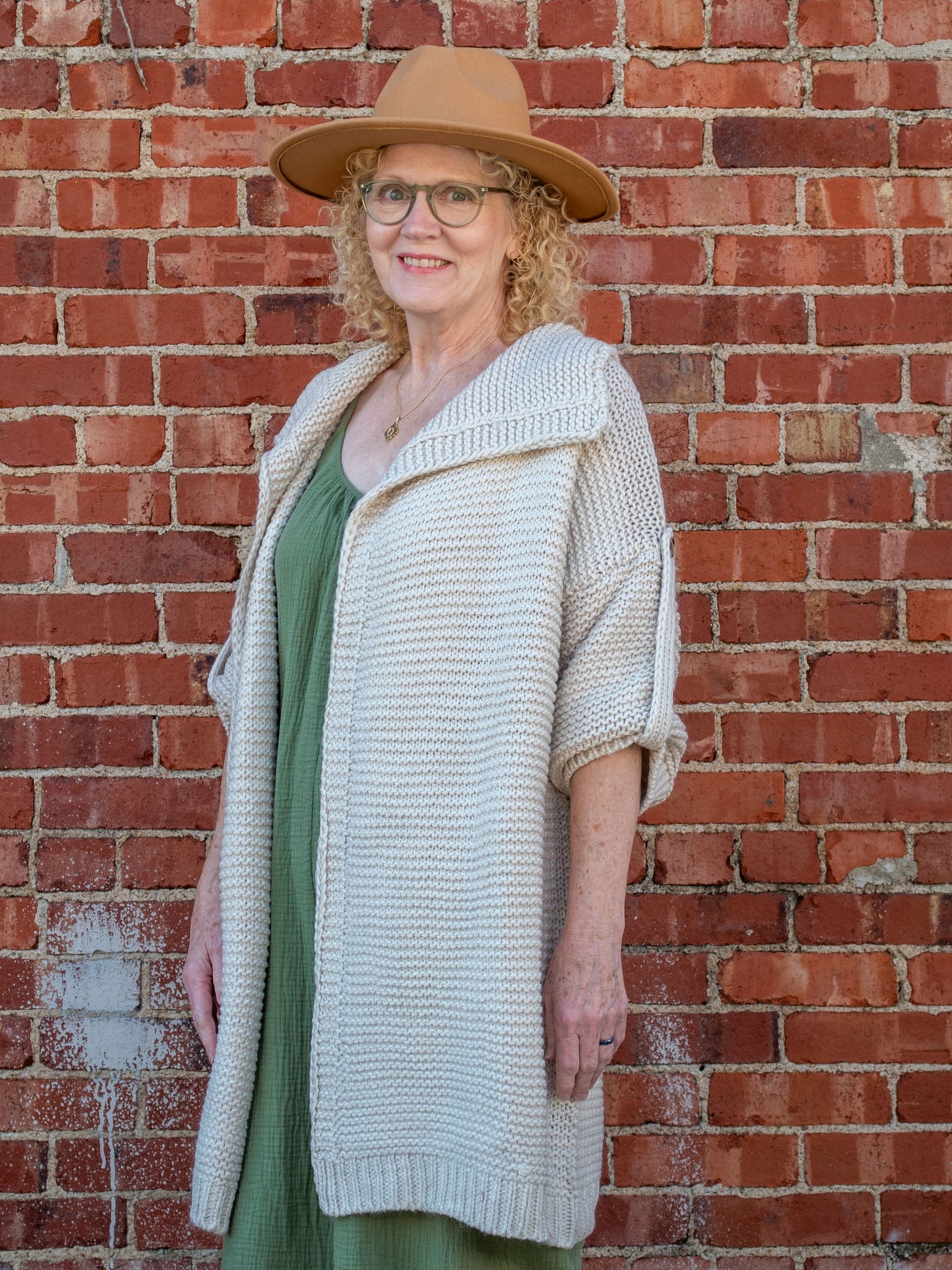 A model wearing a cream, chunky knit cardigan. The model is wearing it over an olive jumpsuit and paired with a brown hat.