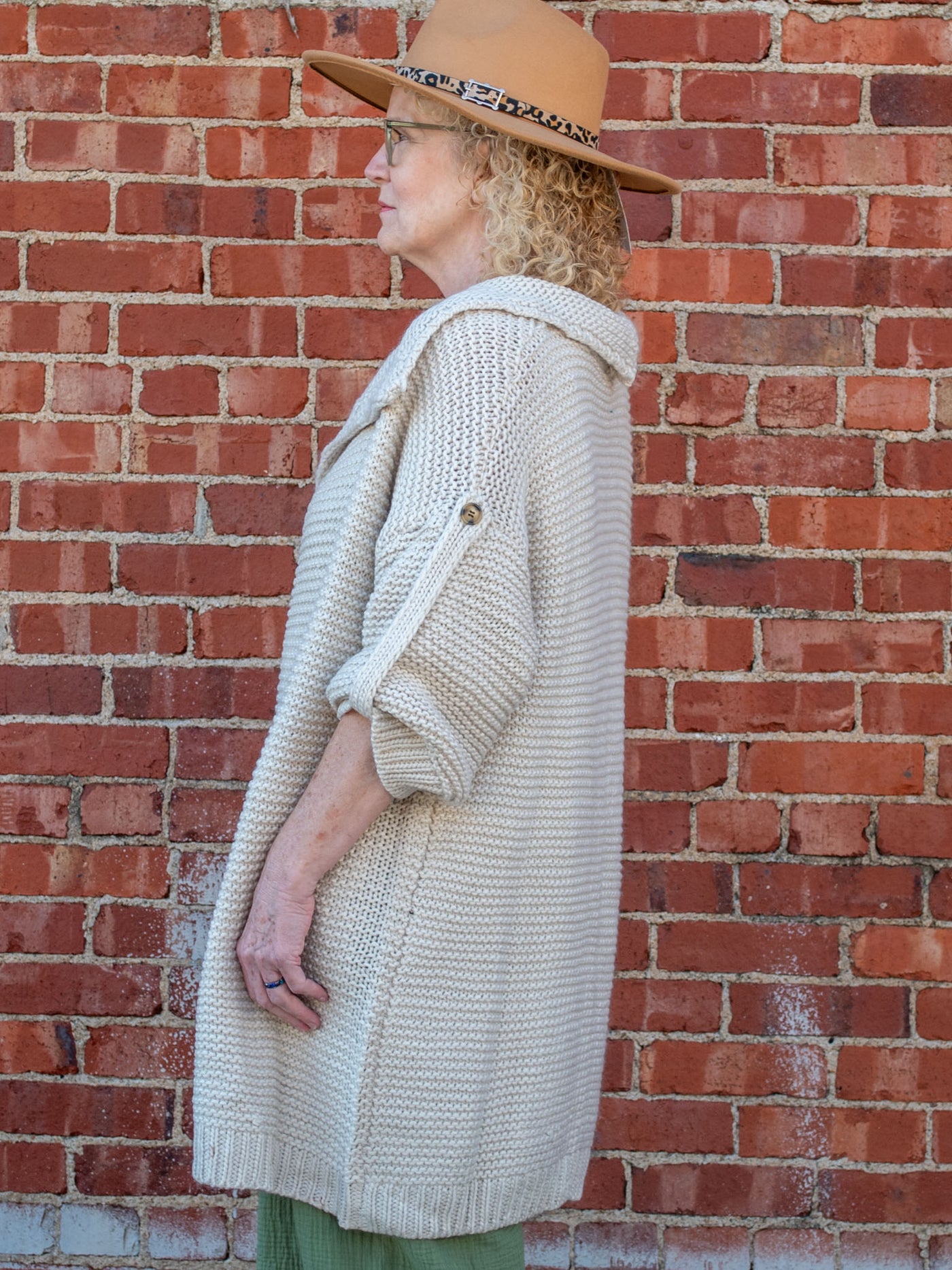 A model wearing a cream, chunky knit cardigan. The model is wearing it over an olive jumpsuit and paired with a brown hat.