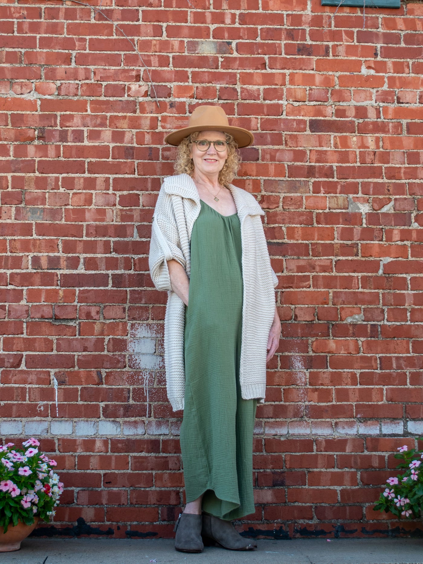 A model wearing a cream, chunky knit cardigan. The model is wearing it over an olive jumpsuit and paired with gray boots and a brown hat.