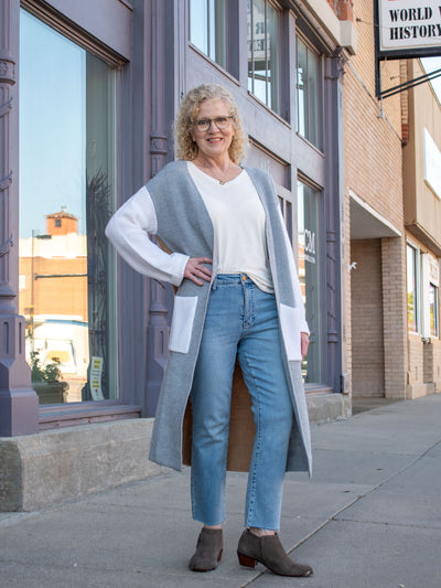 A model wearing a long white, gray and brown color block knit cardigan. The model has it paired with a white top, light wash denim and gray booties.
