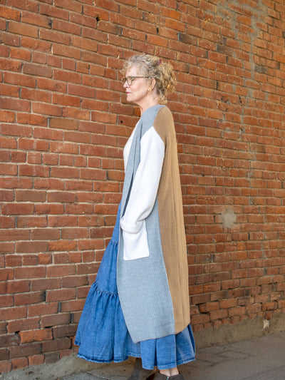 A model wearing a long white, gray and brown color block knit cardigan. The model has it paired with a white top, light wash denim skirt and gray booties.
