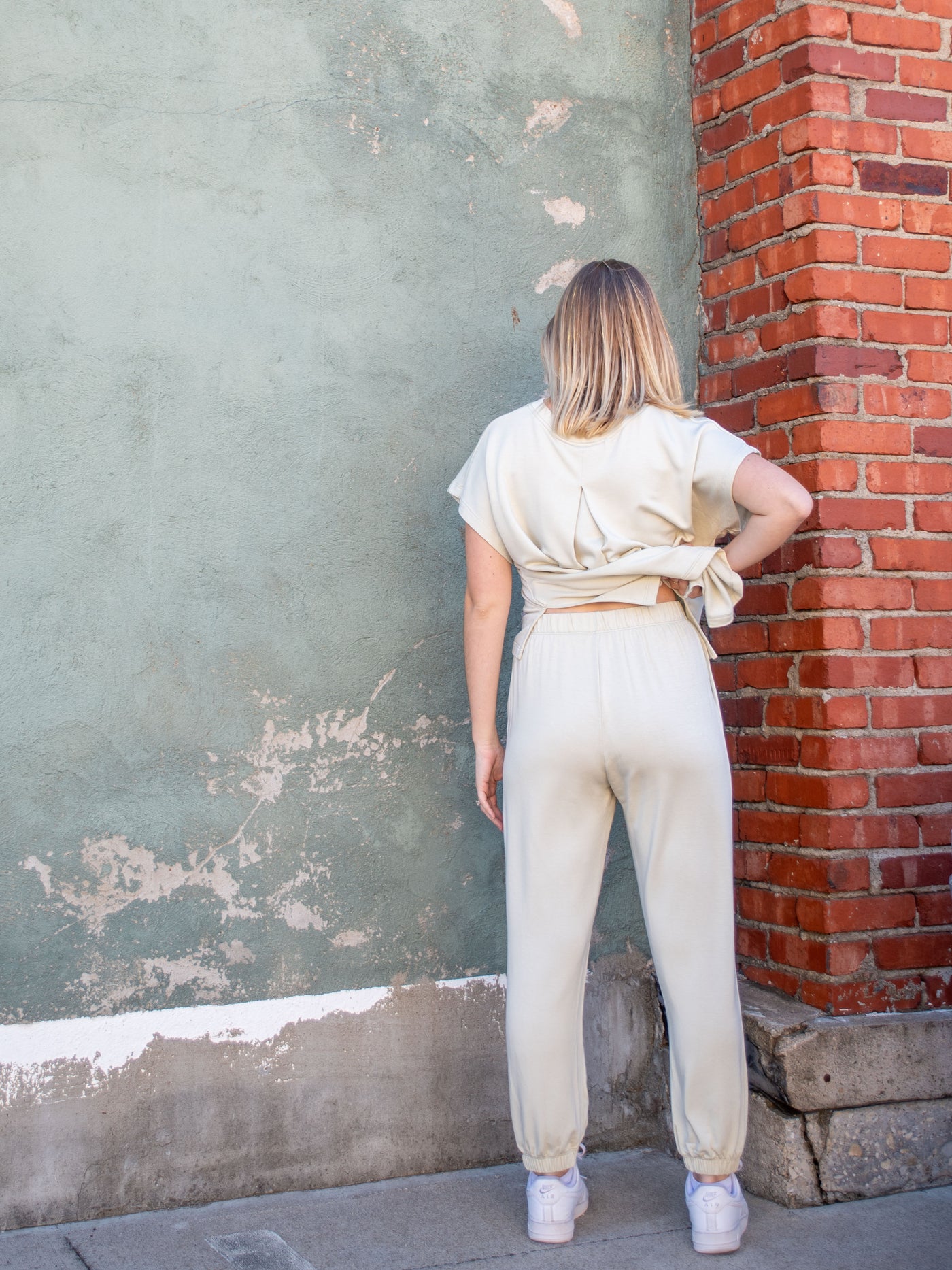 A model wearing an off white jumpsuit with an attached front, detached back, and a jogger pant. The model is wearing it with white sneakers.