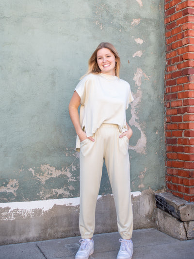 A model wearing an off white jumpsuit with an attached front, detached back, and a jogger pant. The model is wearing it with white sneakers.
