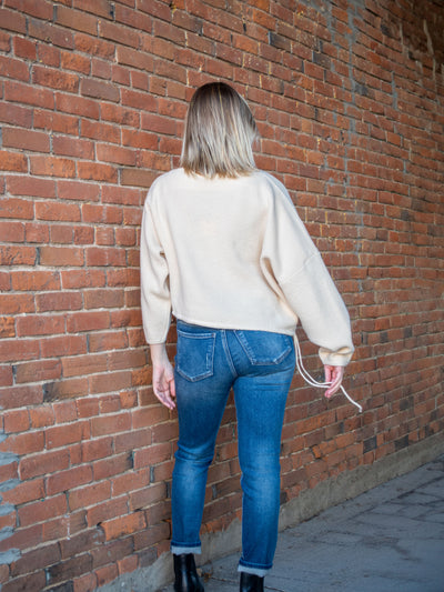 A model wearing a cream, cropped, asymmetrical sweater with an adjustable hem and off the shoulder option. The model has it paired with a medium blue jean and black booties.