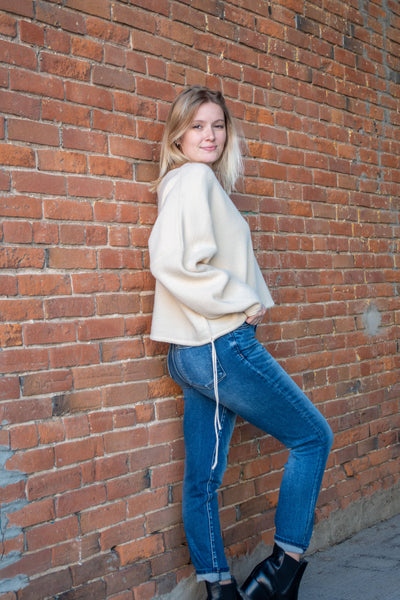 A model wearing a cream, cropped, asymmetrical sweater with an adjustable hem and off the shoulder option. The model has it paired with a medium blue jean and black booties.