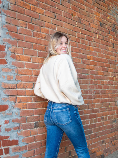 A model wearing a cream, cropped, asymmetrical sweater with an adjustable hem and off the shoulder option. The model has it paired with a medium blue jean.