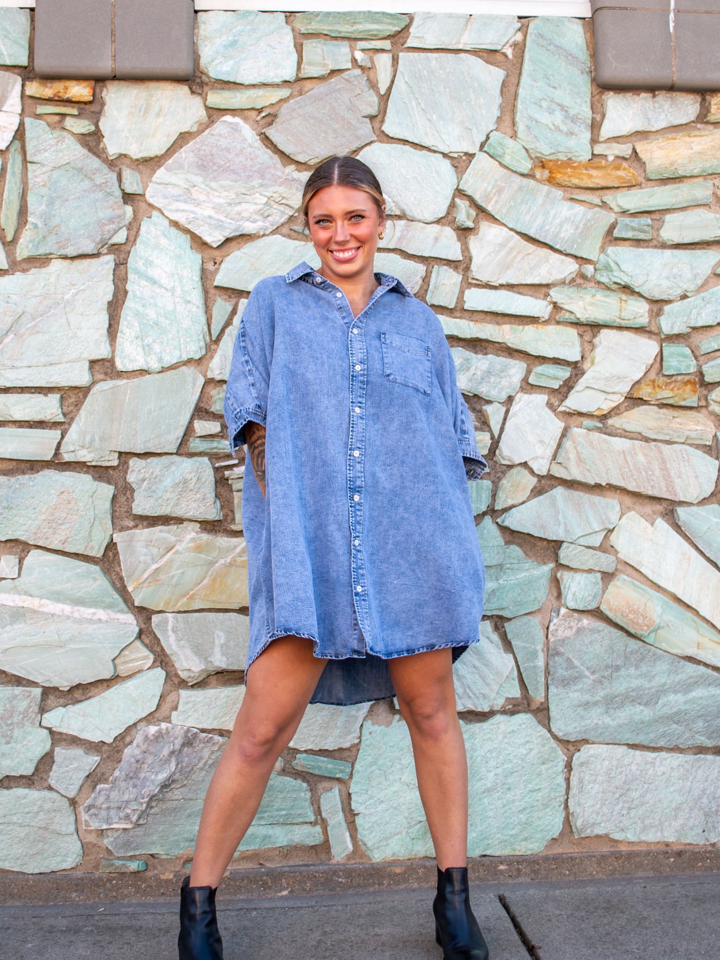 A model wearing a denim wash oversized fit, button up dress with a collar and chest pocket. The model has it paired with black booties.