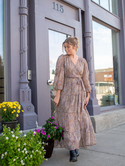 A model wearing a wrap style maxi dress with a light brown colored paisley print all over it. The model has it paired with black booties.