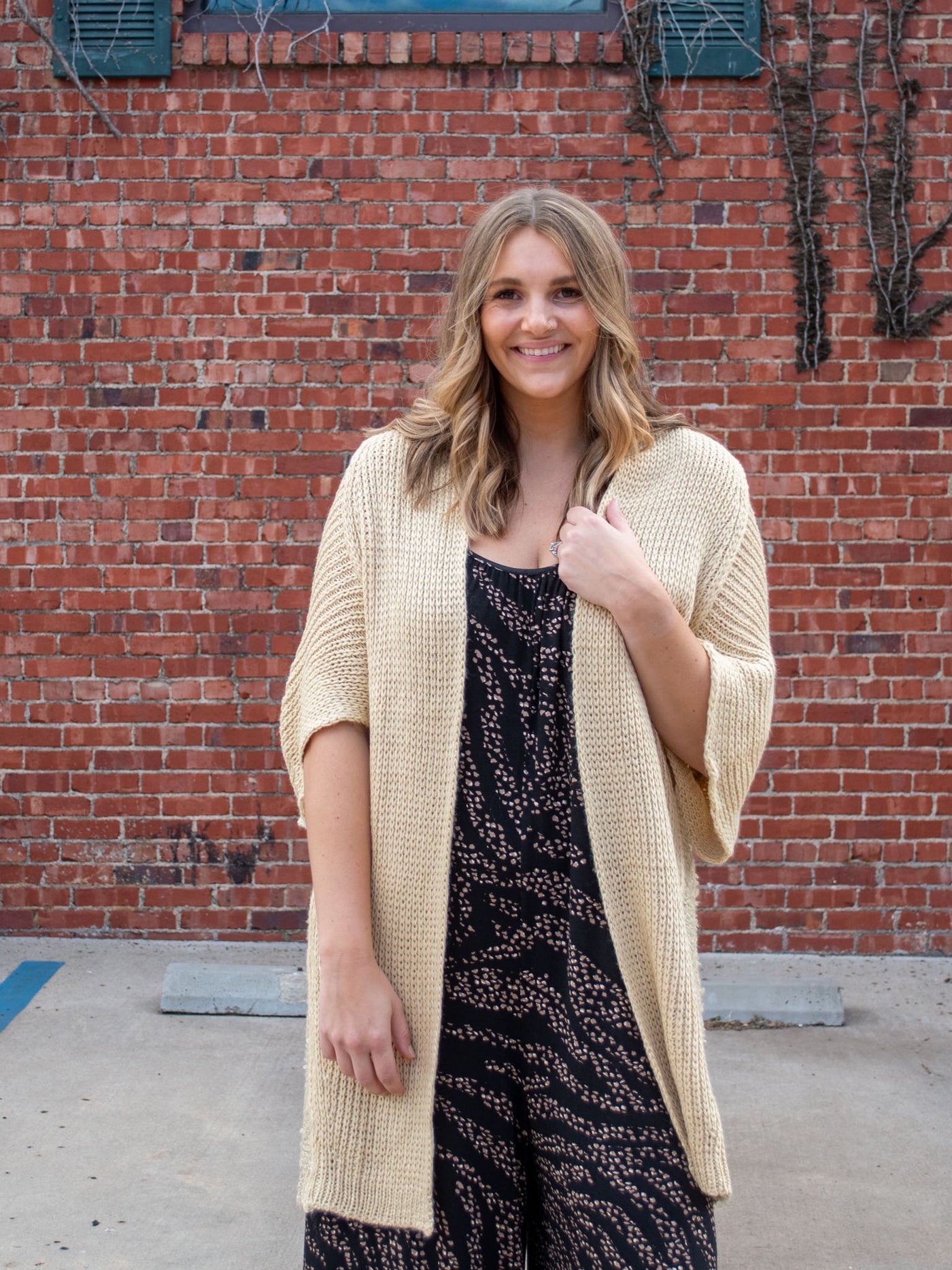 A model wearing a tan colored oversized chunky cardigan. The model has it paired over a black animal print jumpsuit. 