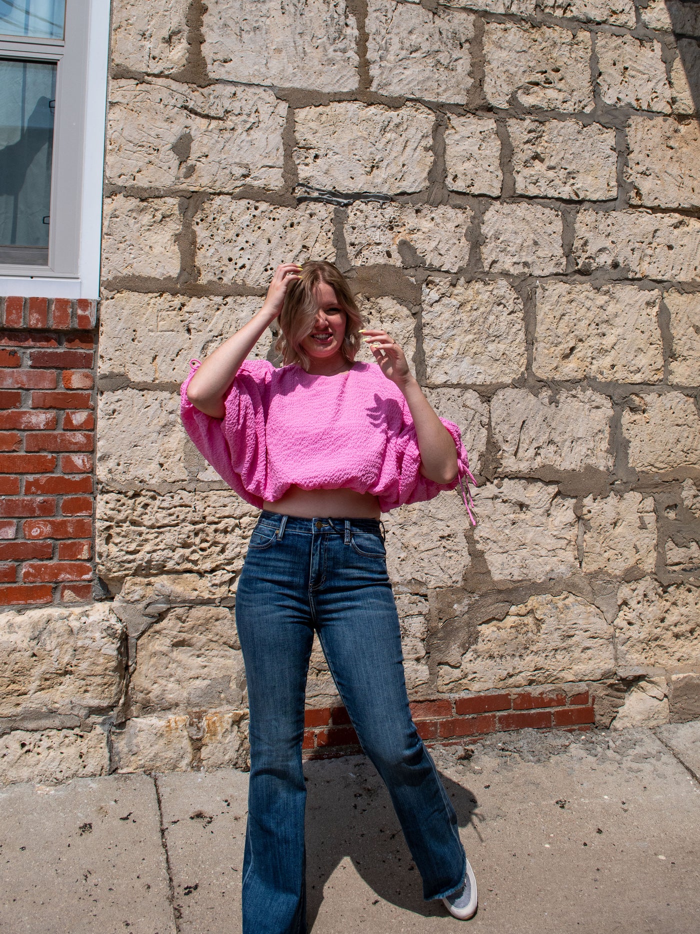 A model wearing a pink voluminous drawstring crop top with a textured fabric. She has it on with flare jeans and sneakers.