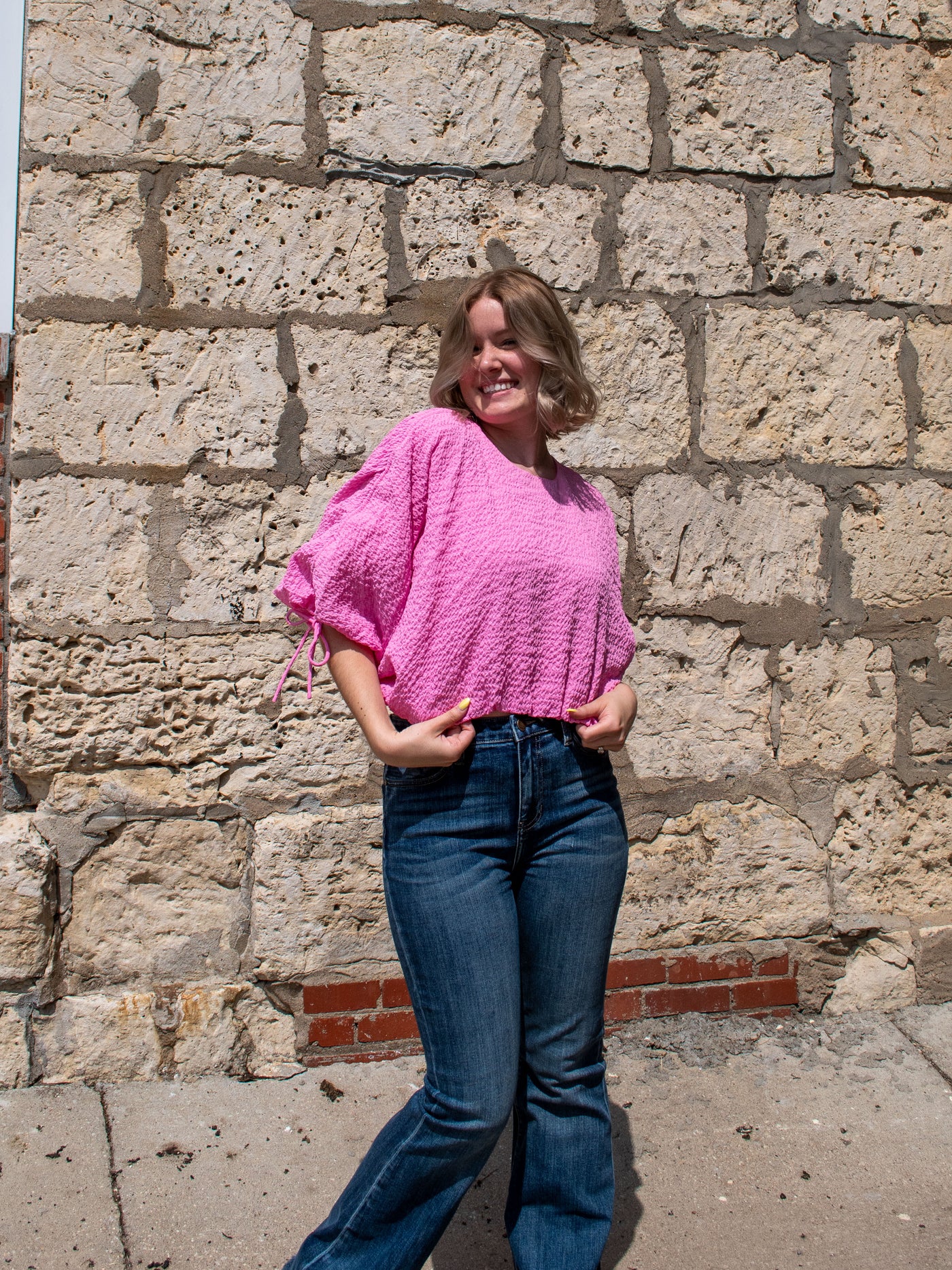 A model wearing a pink voluminous drawstring crop top with a textured fabric. She has it on with flare jeans and sneakers.