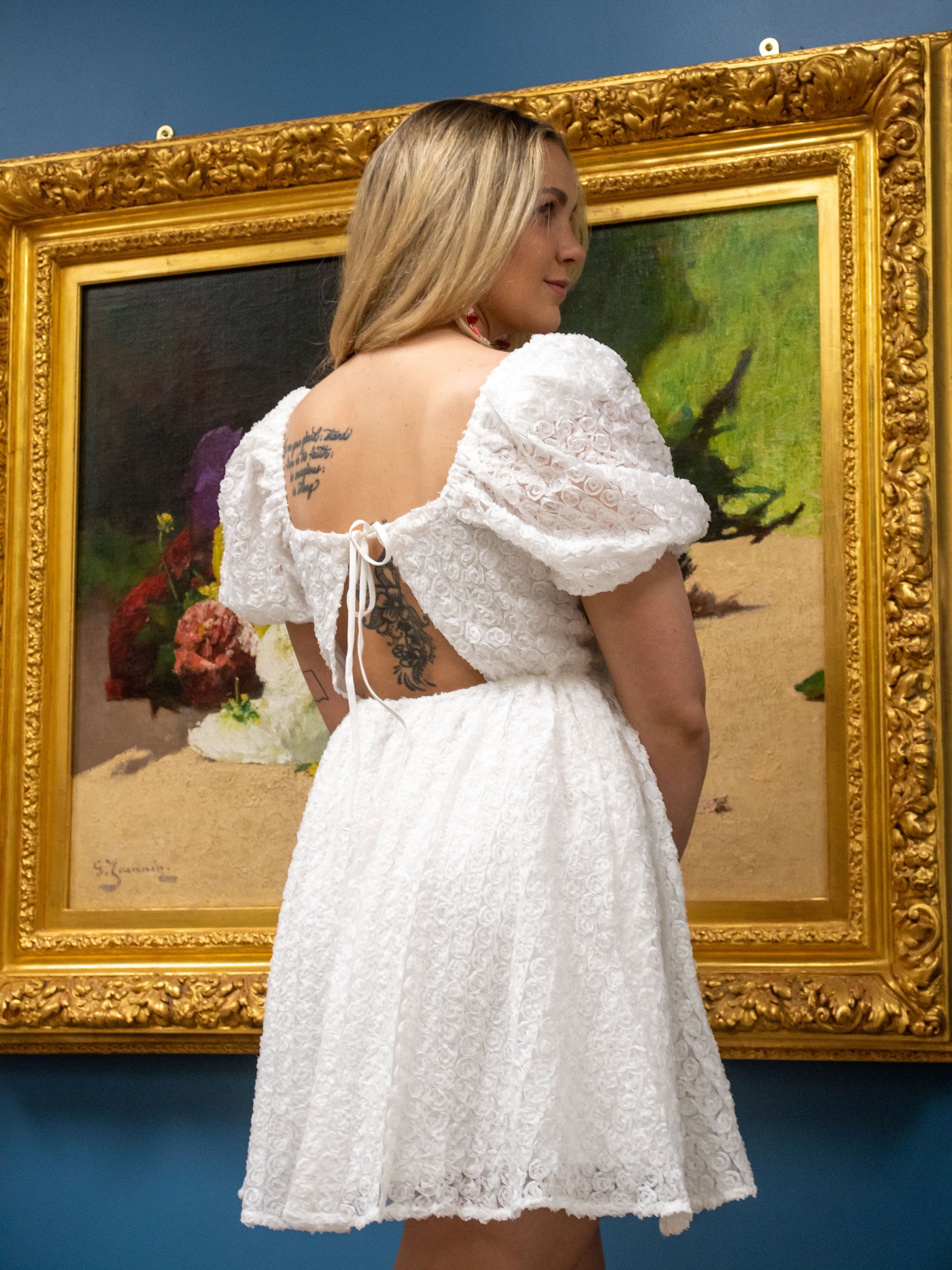 A model wearing a white lace floral mini dress with puff sleeves and a tie back detail. The model has it on with light pink boots.