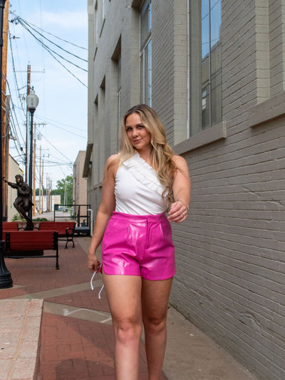 A model wearing a white asymmetrical ruffle bodysuit with hot pink pleather shorts and pink heels.