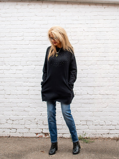 A model wearing a long black sweater top with sparkle around the top. The model has it paired with a medium wash jean.