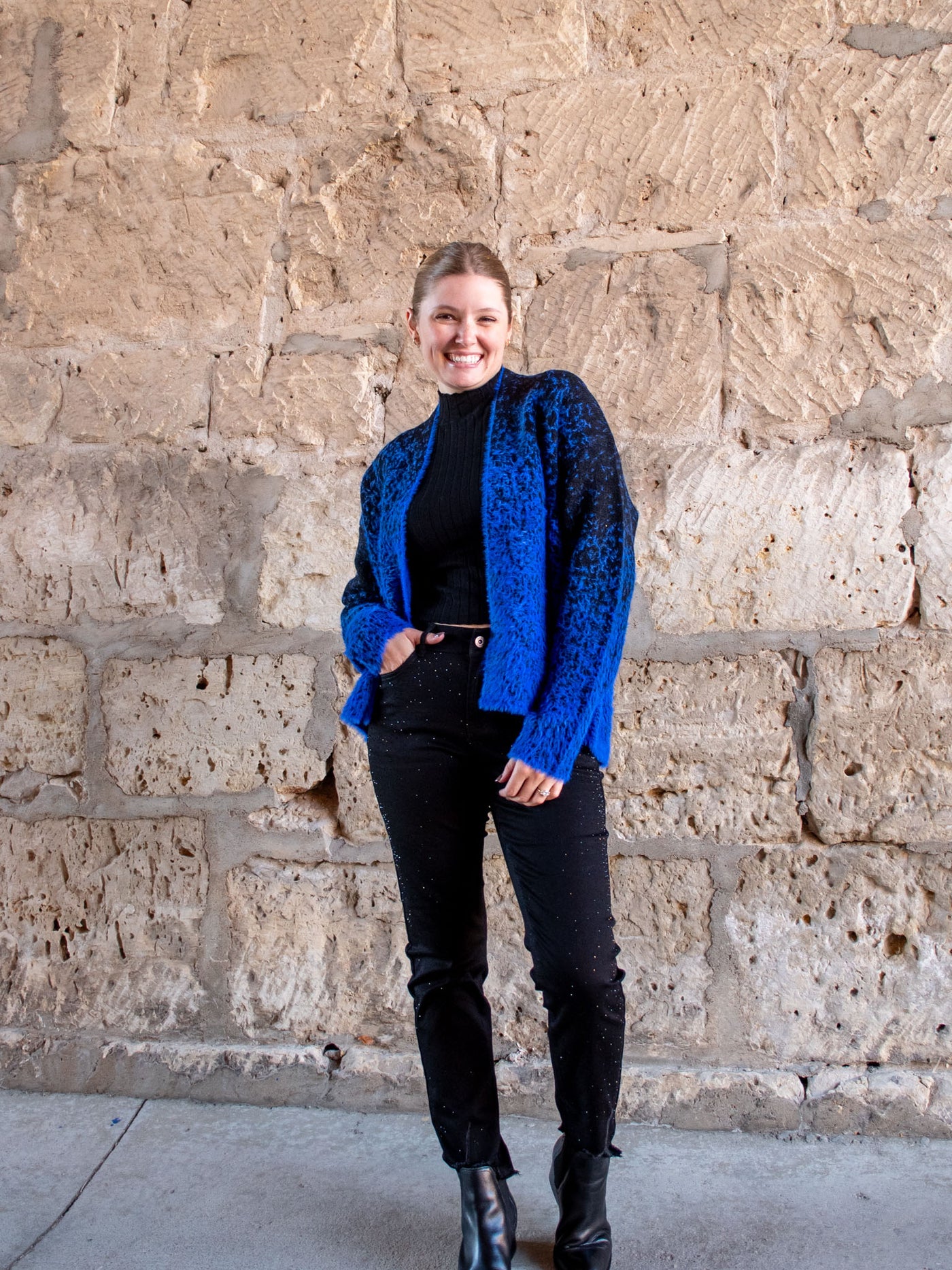 A model wearing a blue and black fuzzy open front cardigan. The model has it over a black mock neck, black jeans, and black booties.