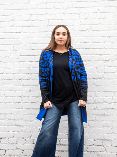 A model wearing a black and blue open front, animal print cardigan. The model has it paired over a black sweater and wide leg, dark wash jeans.