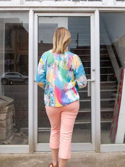 A model wearing a blue colorful patterned, short, knit cardigan. The model has it paired with a white tank and peach pants.